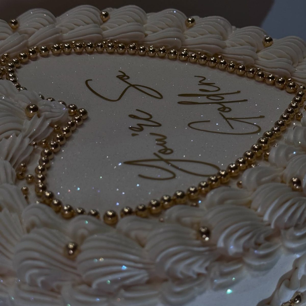 Heart-shaped white cake with gold pearl borders and gold cursive lettering.
