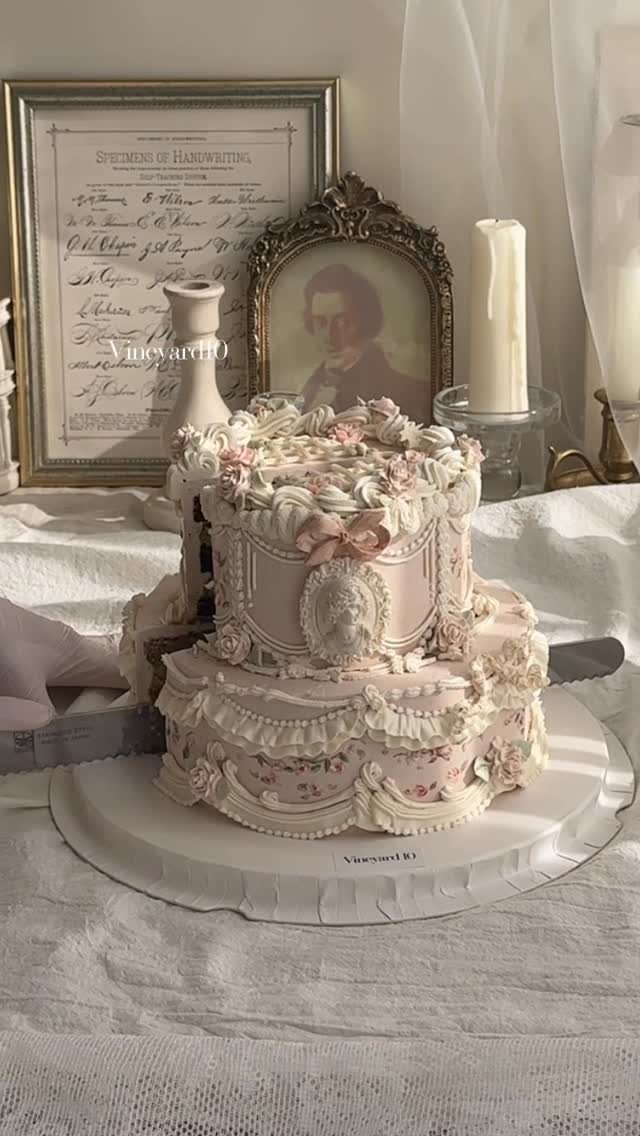 Two-tier pink vintage cake with ornate frosting ruffles and floral decorations on linen.