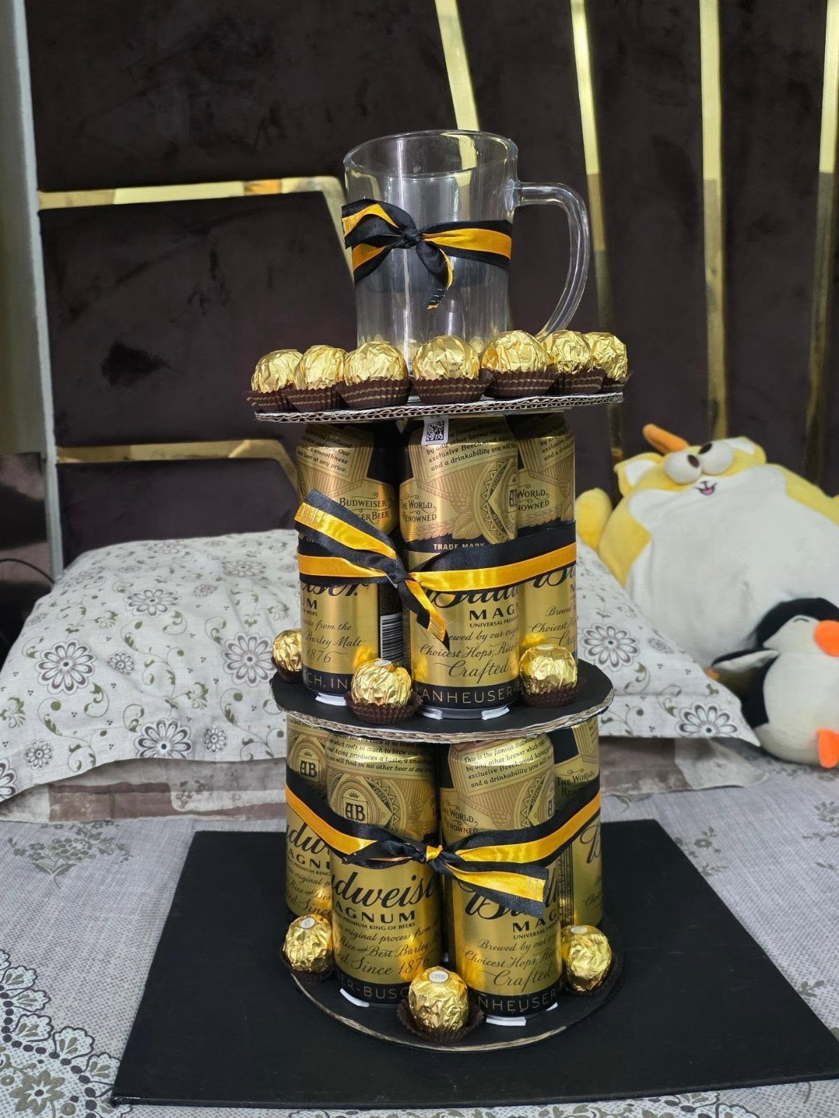 Tiered beer can cake with gold Budweiser cans, chocolates, and a glass mug.
