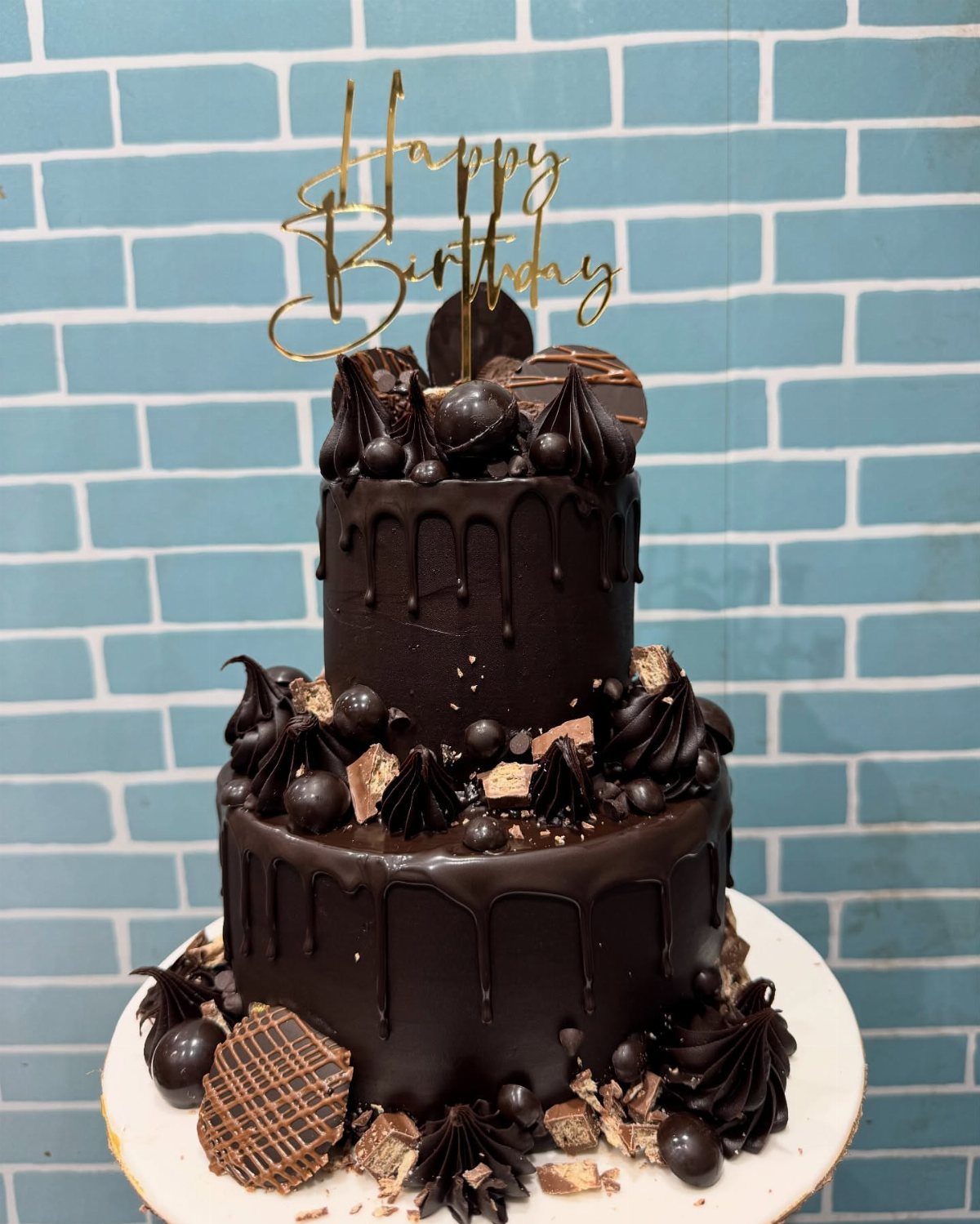 Two-tier chocolate cake with dark frosting and a gold Happy Birthday topper.