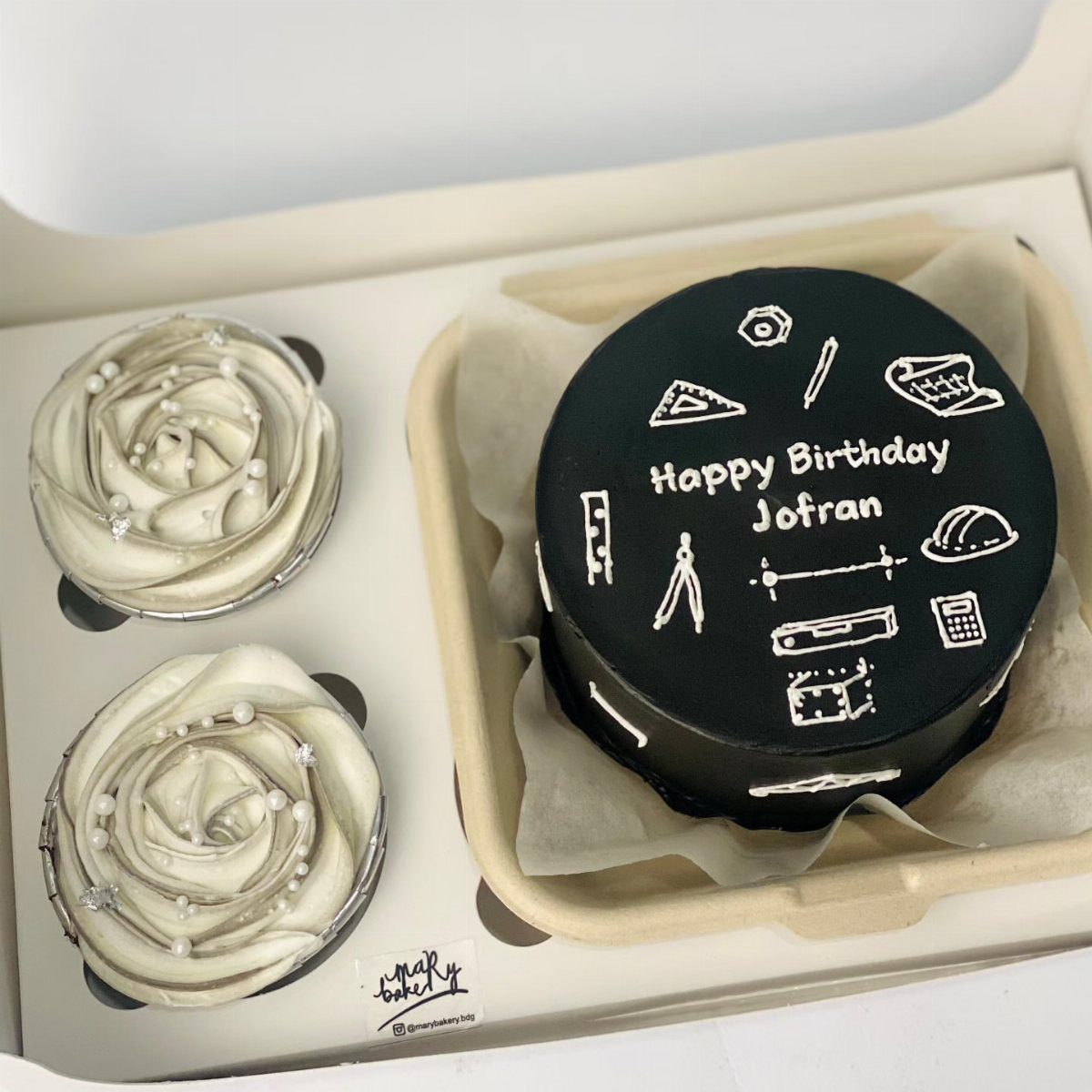 Black fondant birthday cake with white architectural drawings and two white cupcakes.