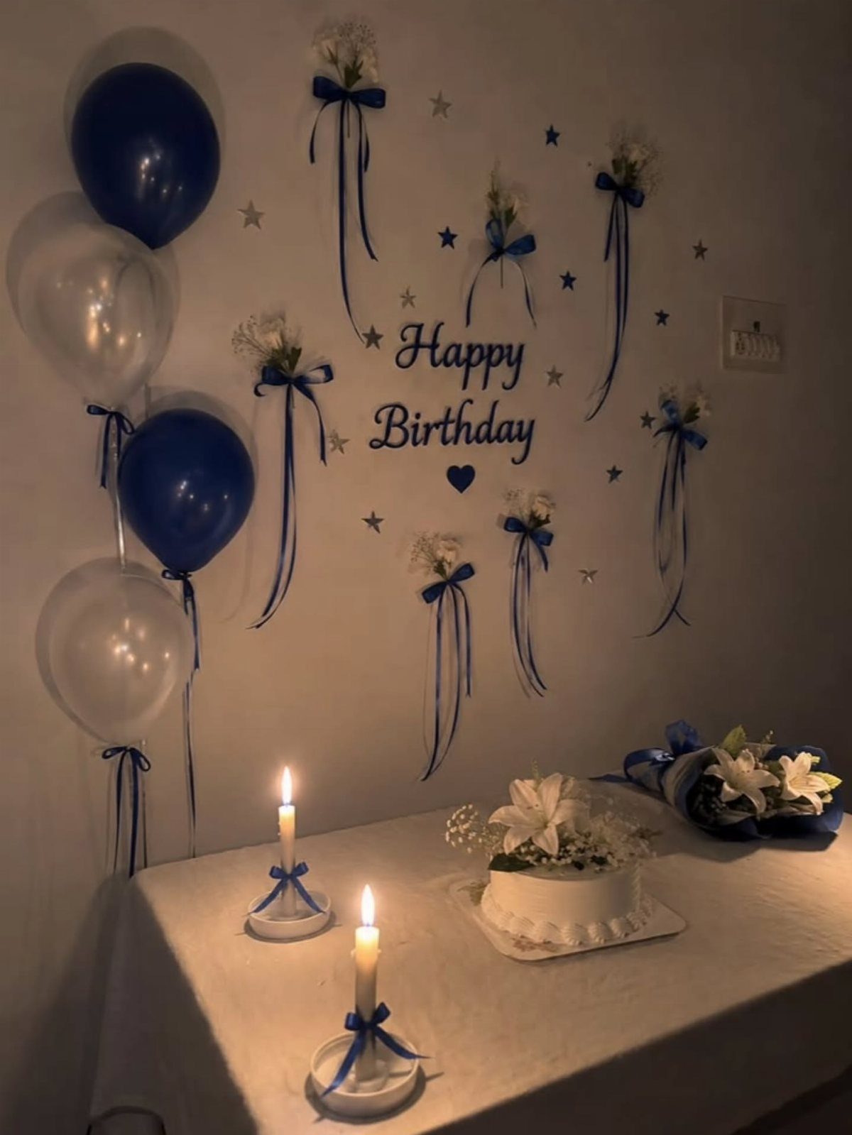 Birthday decorations featuring blue balloons, a white cake, and ribbons on a wall.