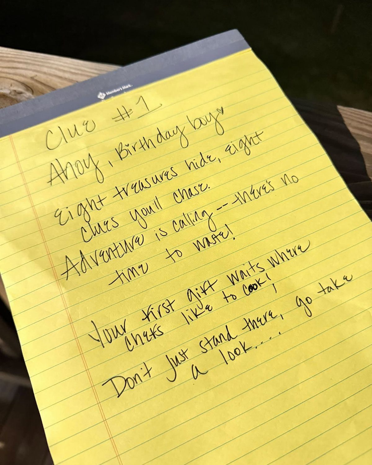 Handwritten scavenger hunt clue on yellow lined paper resting on a wooden surface.
