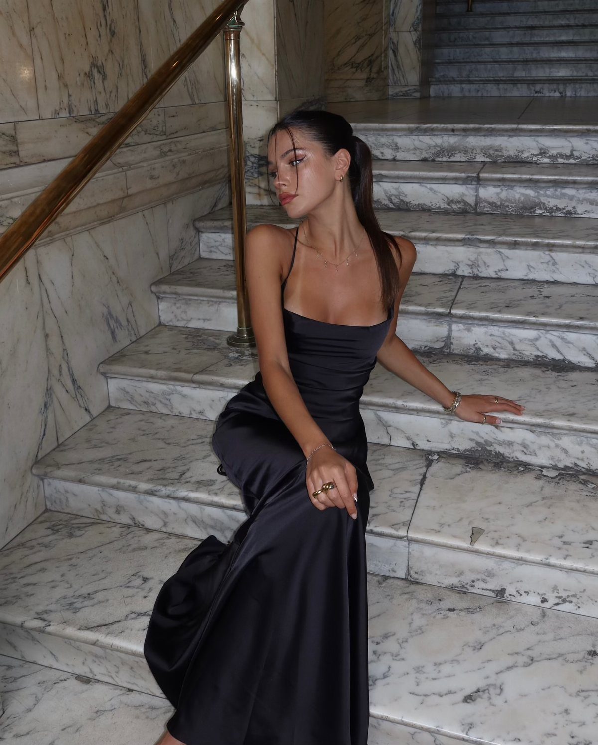 Woman in a long black satin slip dress sitting on white marble stairs.
