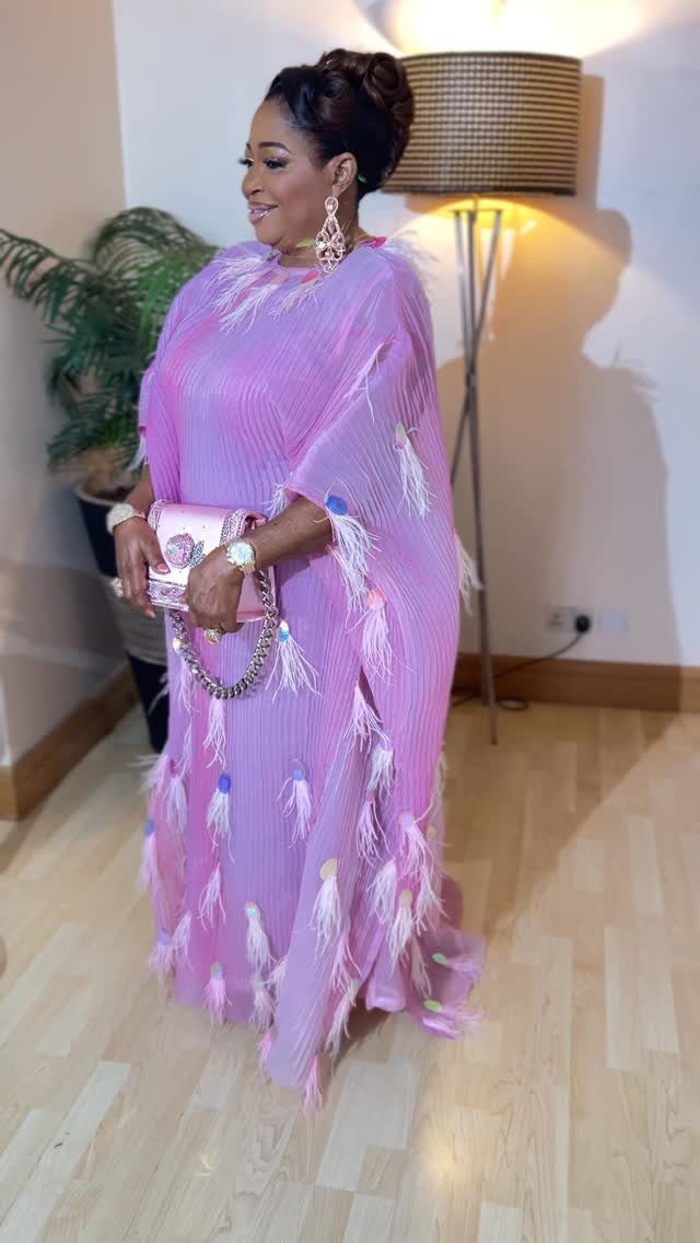 Woman in a purple pleated gown with tassels holding a pink handbag indoors.