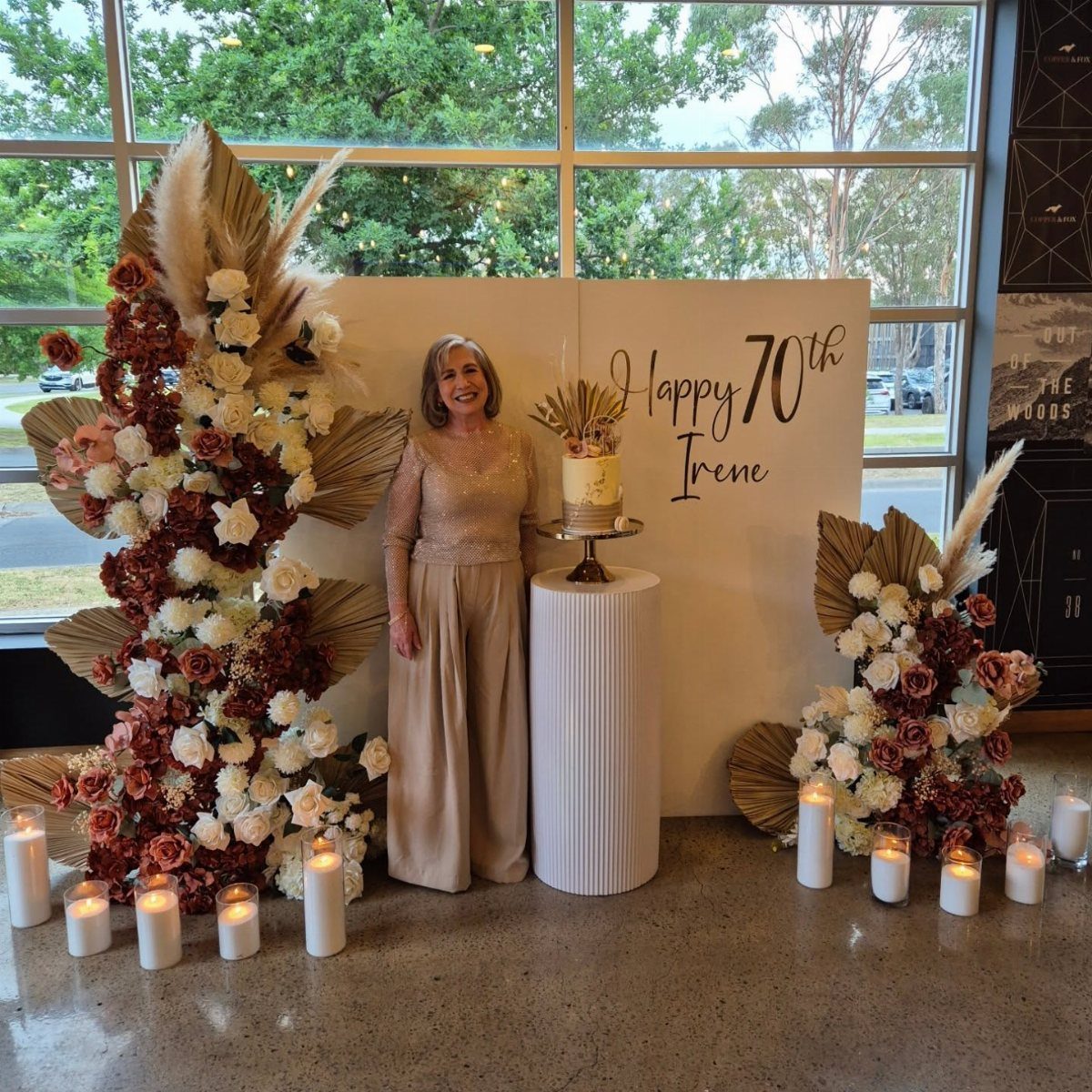 Woman in champagne lace outfit standing by 70th birthday cake and floral decor