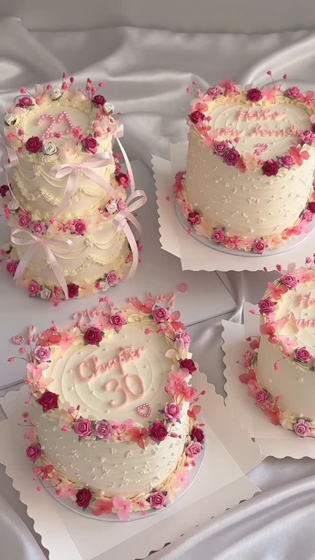 White frosted birthday cakes decorated with pink flowers and ribbons on white fabric.