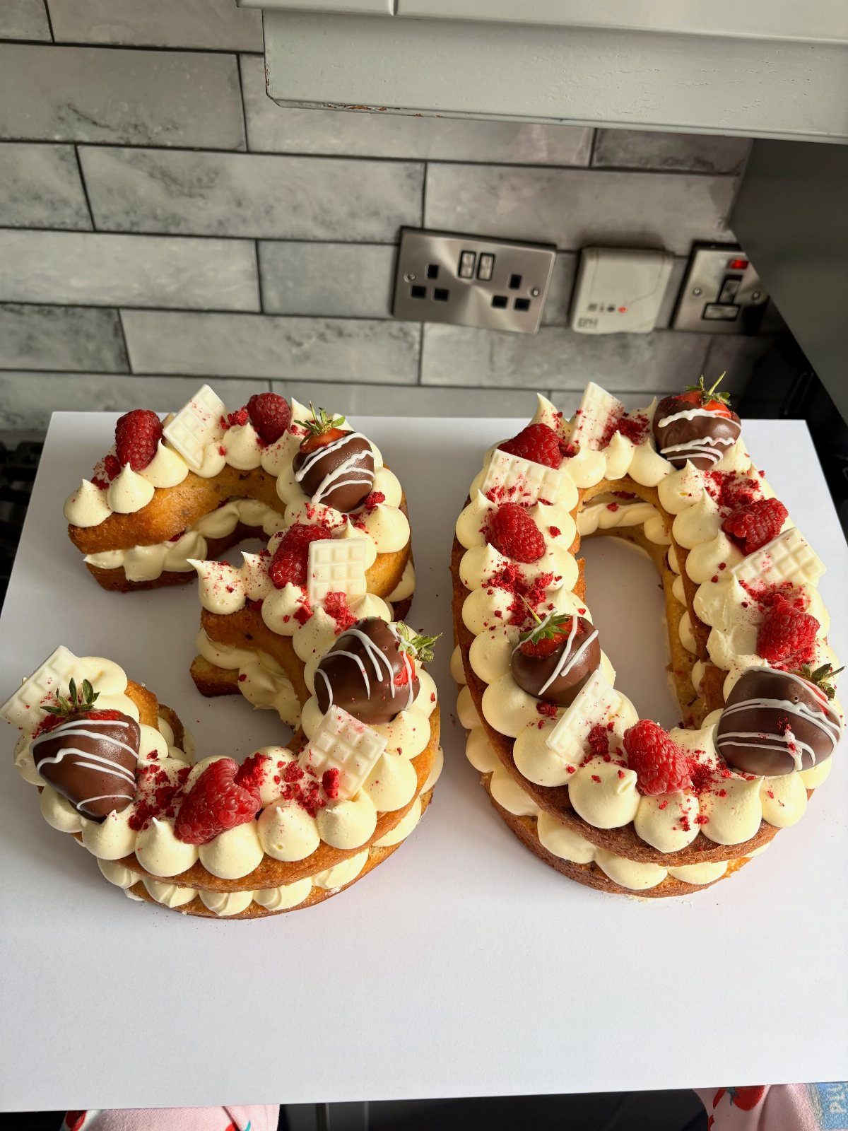 Number 30 cake decorated with cream, raspberries, and chocolate-dipped strawberries on white background.