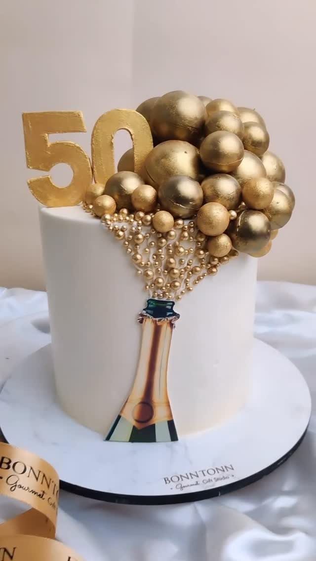 White birthday cake with gold bubble decorations, a gold 50 topper, and champagne bottle.