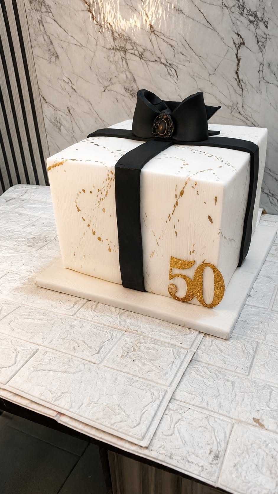 Square white gift-shaped cake with black ribbon, gold accents, and gold number 50.
