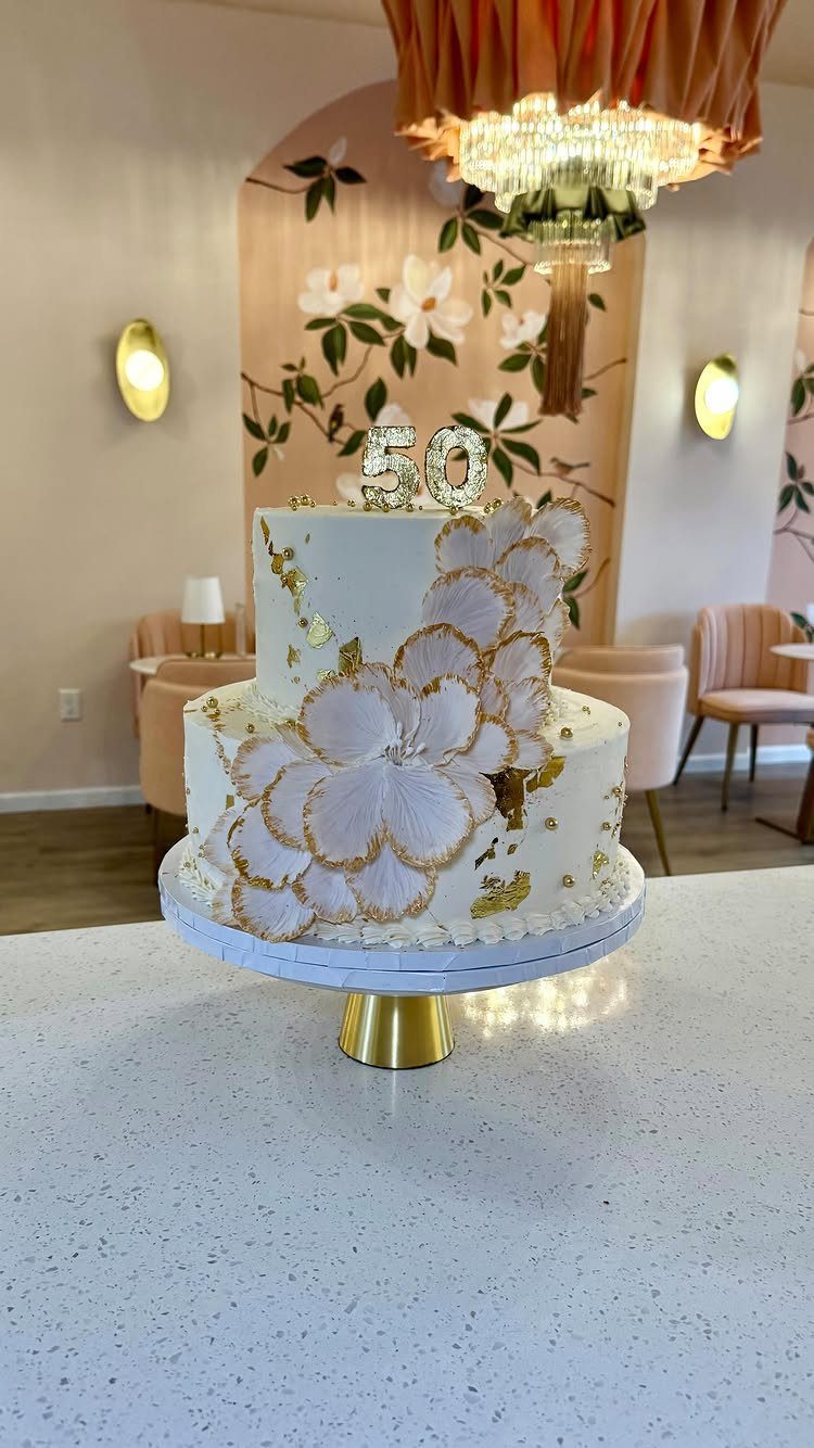 Two-tier white cake with gold leaf accents, white flowers, and a gold 50 topper.