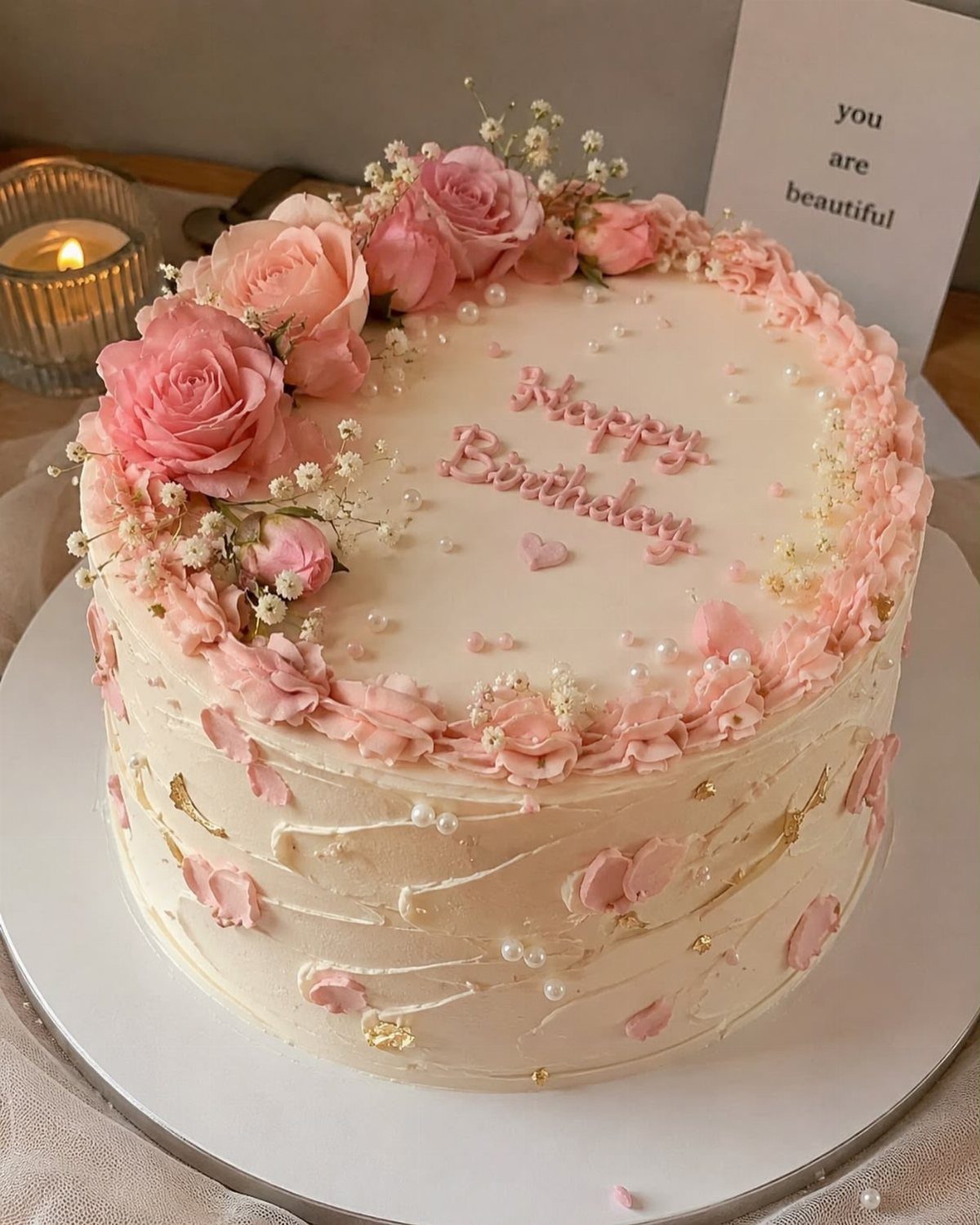 White frosted cake decorated with pink roses, buttercream swirls, and gold accents.