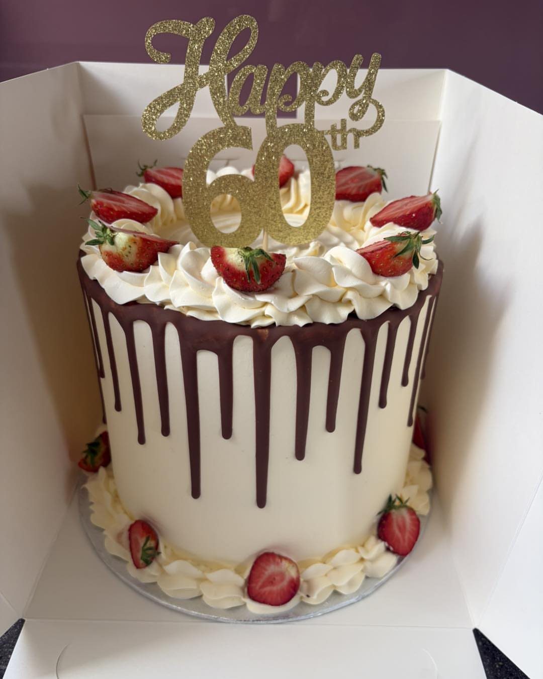 Tall white cake with chocolate ganache drips, fresh strawberries, and gold 60th topper.