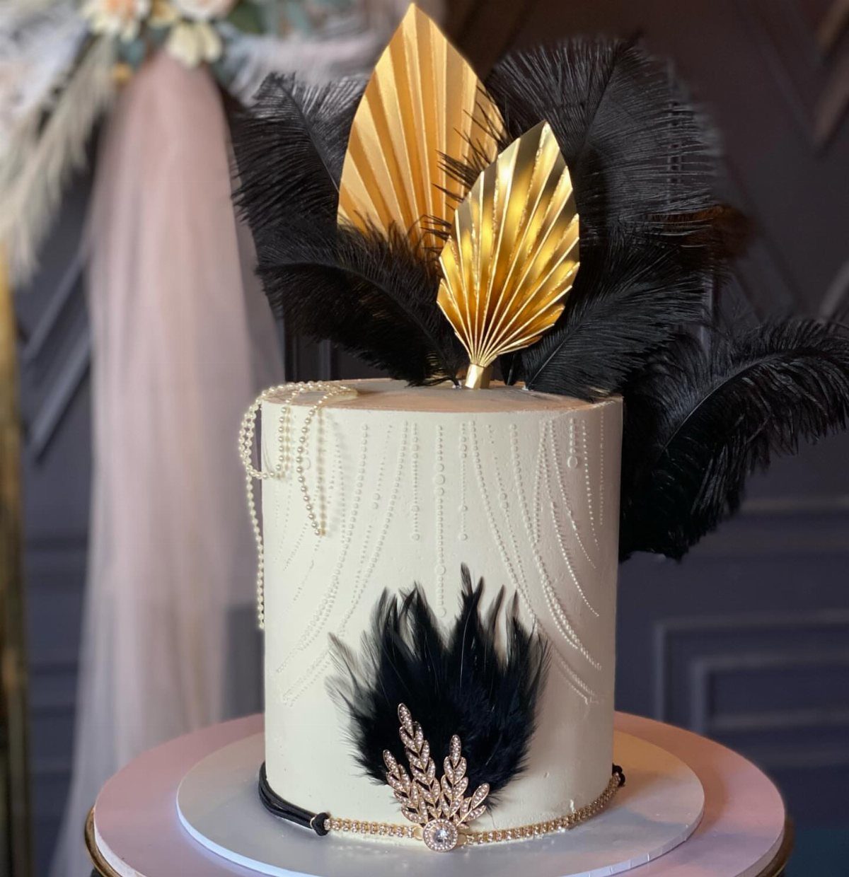 White cylindrical cake decorated with black feathers, gold leaf accents, and pearl beading.