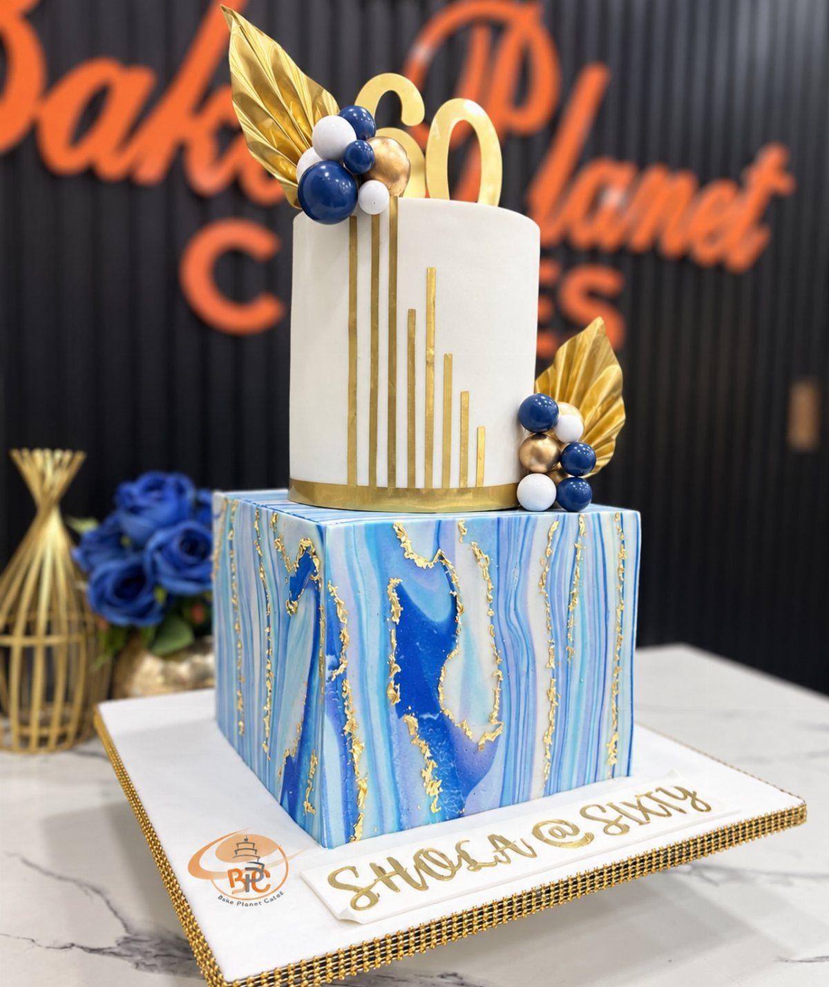Two-tier birthday cake with blue marble square base and white cylindrical top layer.