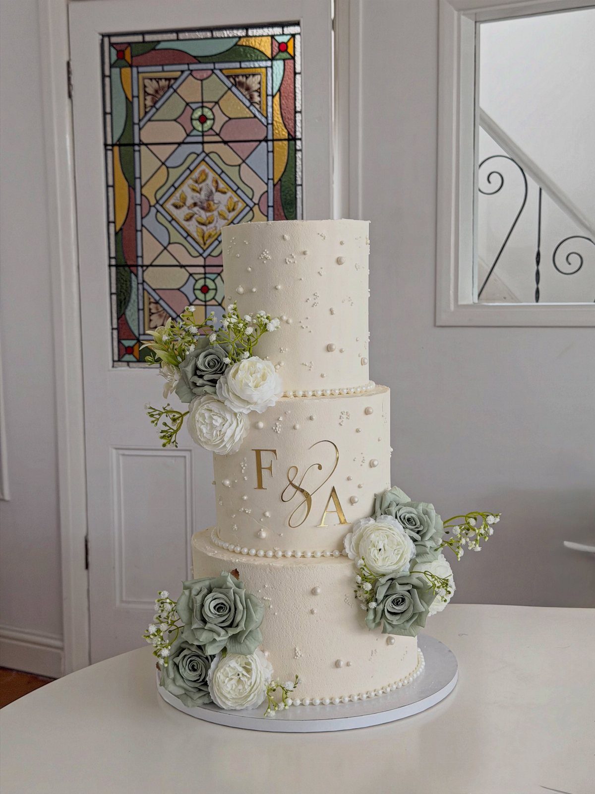 Three-tiered white cake with gold monogram, white pearls, and sage green roses.