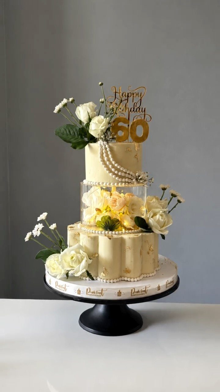 Tiered cream birthday cake with edible pearls, white roses, and gold 60th topper.