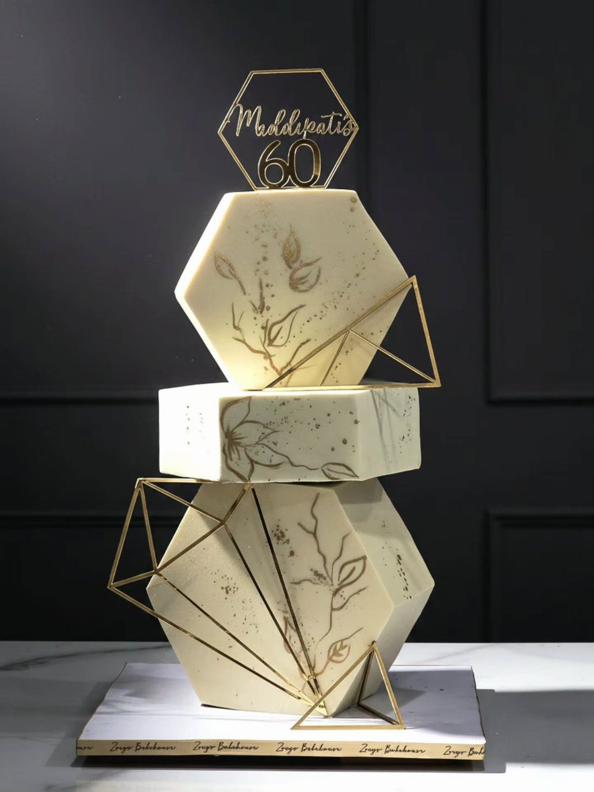Three-tiered white hexagonal cake with gold geometric accents and a 60th birthday topper.