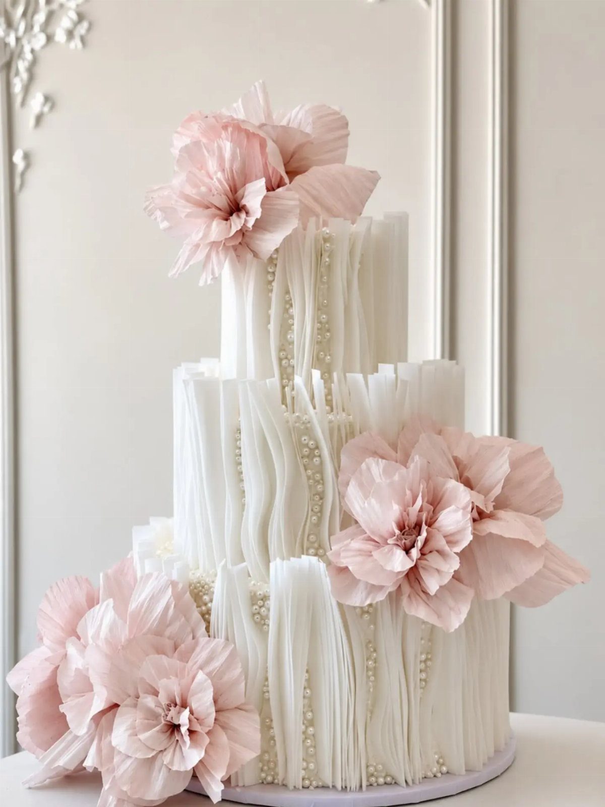 Three-tiered white pleated cake decorated with large pale pink flowers and white pearls.