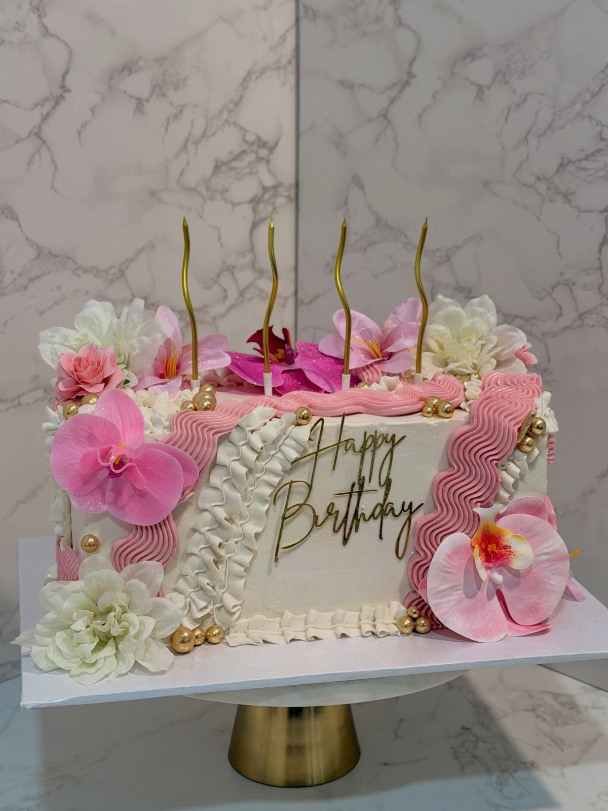 White rectangular birthday cake with pink ruffles, orchids, and gold Happy Birthday topper.