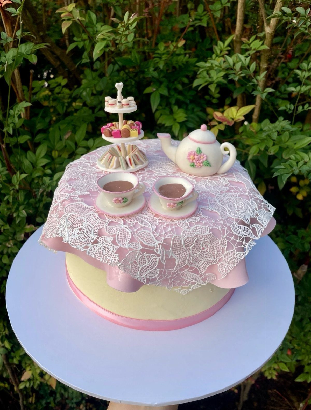 Tea party cake with pink lace, teapot, teacups, and macarons in a garden