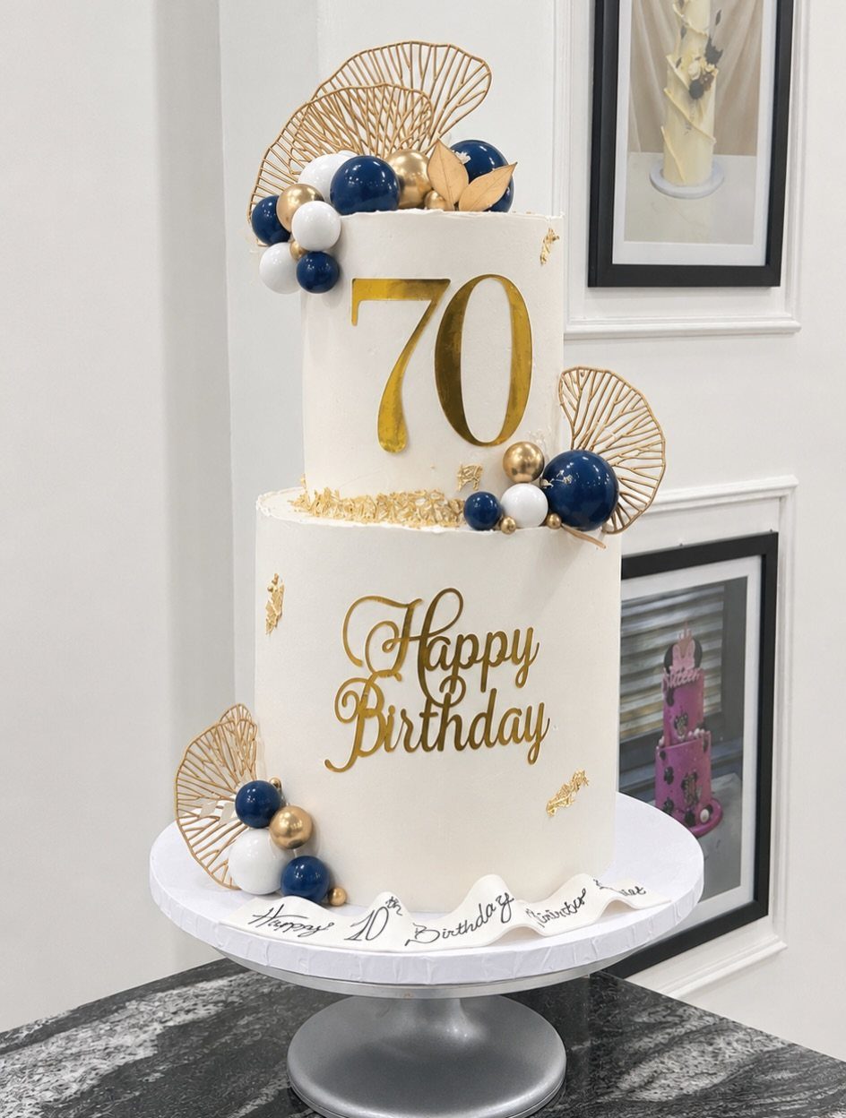 Two-tier white 70th birthday cake with gold lettering and navy blue sphere decorations.