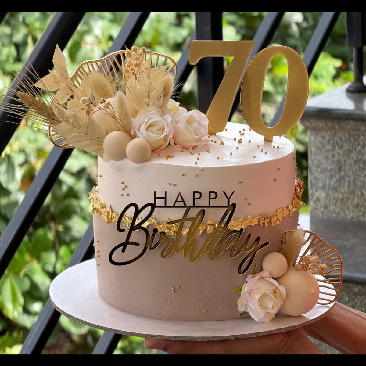 White 70th birthday cake decorated with gold accents, pale pink roses, and pampas grass.