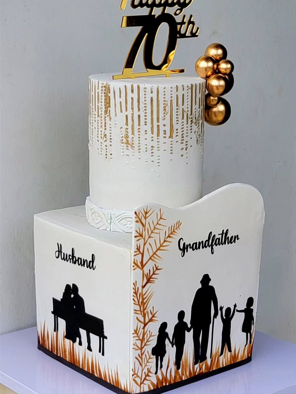 Two-tier white 70th birthday cake with gold accents and husband and grandfather silhouettes