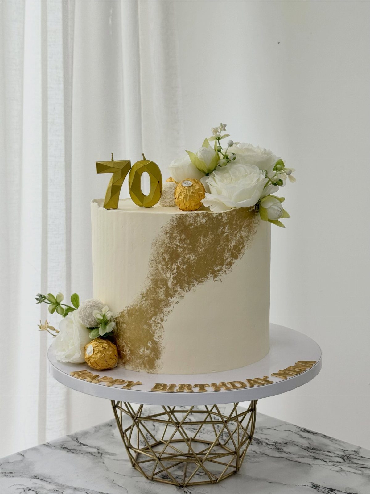 White birthday cake with gold accents, white roses, and gold number 70 candles.