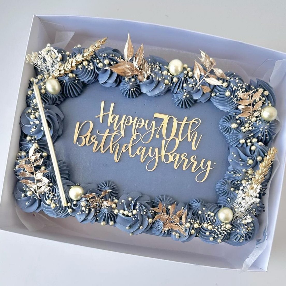 Blue-grey rectangular cake with gold leaves, pearls, and Happy 70th Birthday topper.