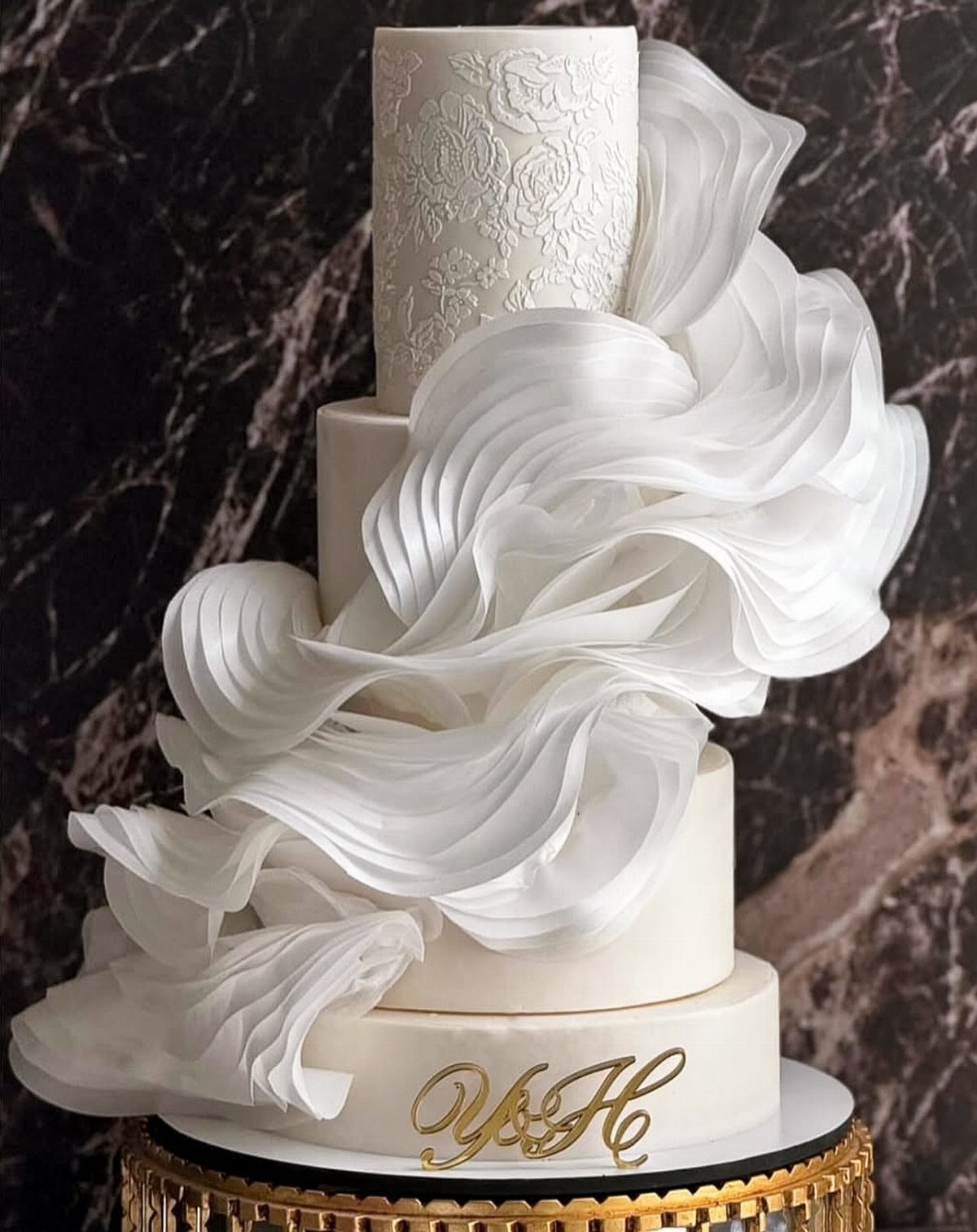 Multi-tiered white cake with flowing ruffles, lace patterns, and gold monogram initials.