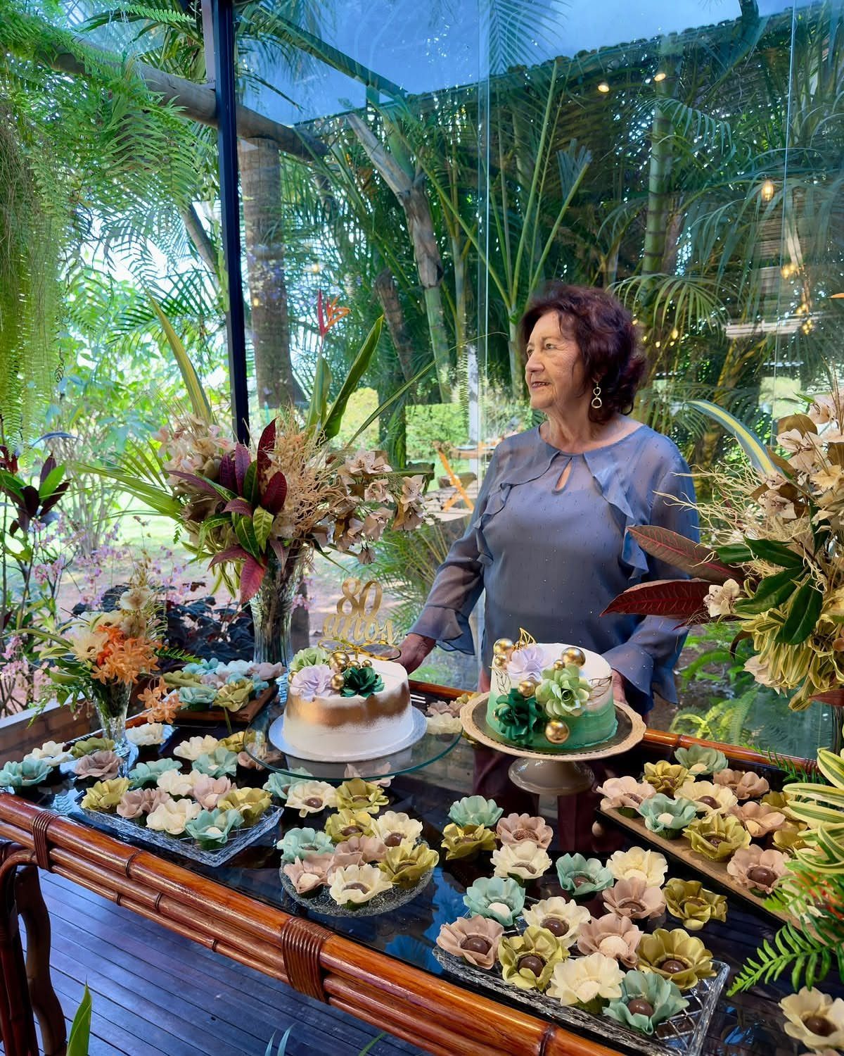 Woman standing behind a dessert table with floral cupcakes and cakes in a sunroom.