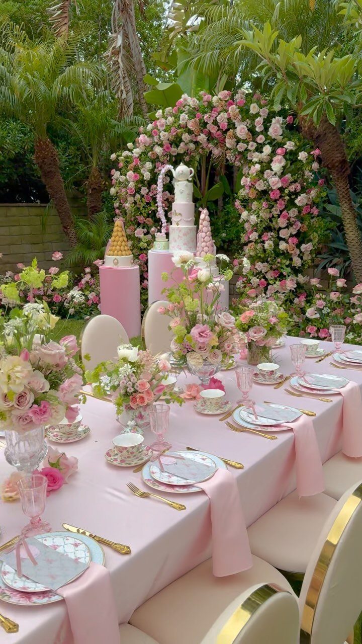 Pink garden party table with floral centerpieces, tea sets, and a floral arch background.