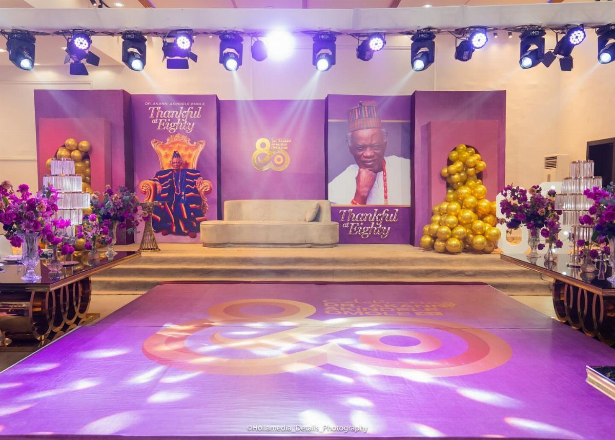 Purple and gold event stage with a portrait, throne, and gold balloon arches.