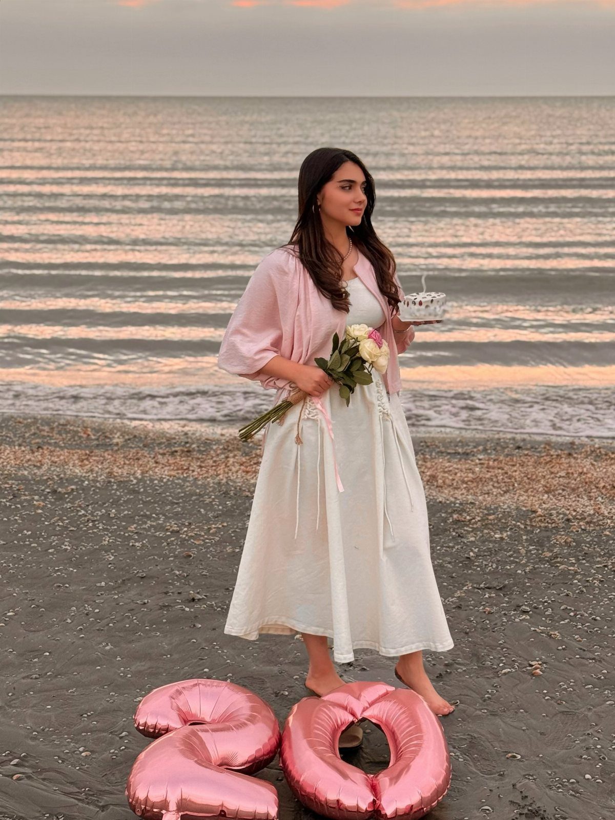 Woman in white dress holding cake and flowers on a beach with number balloons.