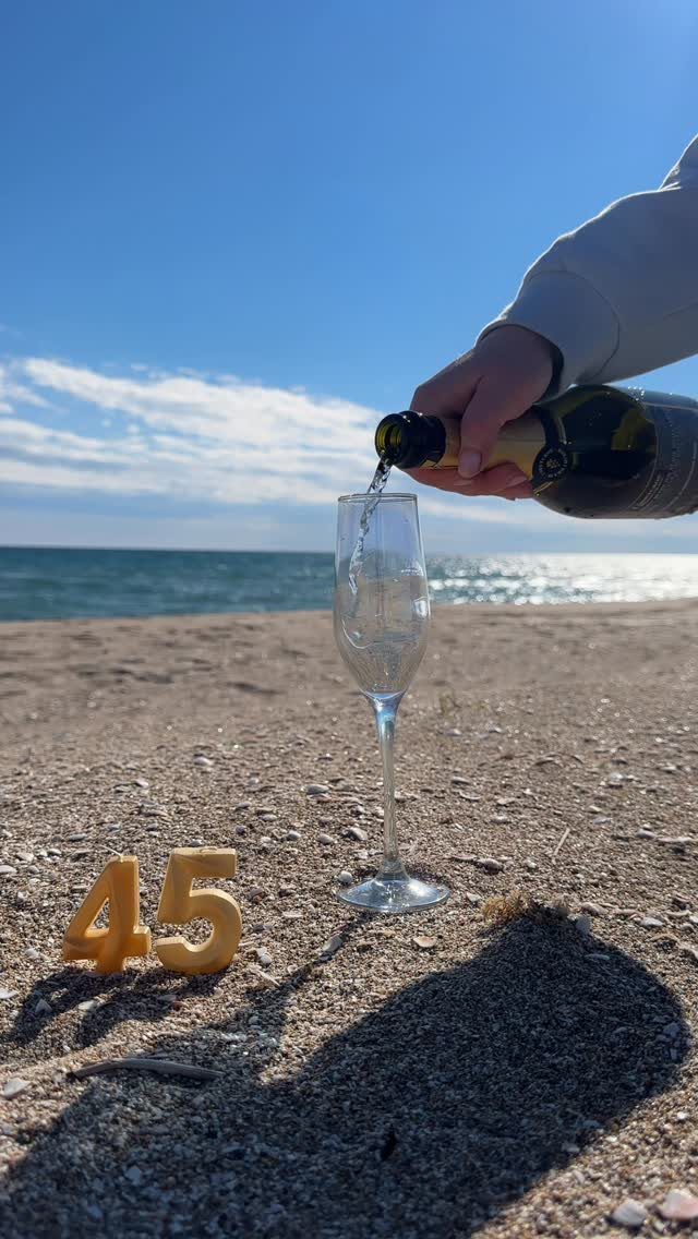 Champagne being poured into a glass on a beach next to number 45 candles.
