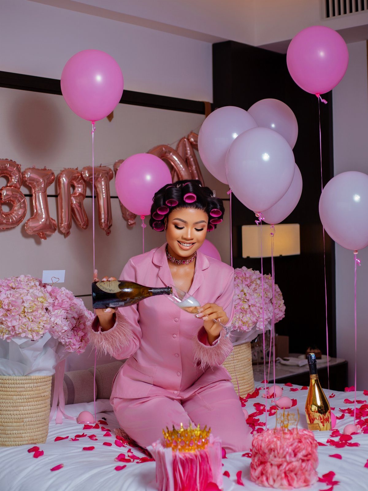 Woman in pink pajamas pouring champagne surrounded by balloons and rose petals indoors