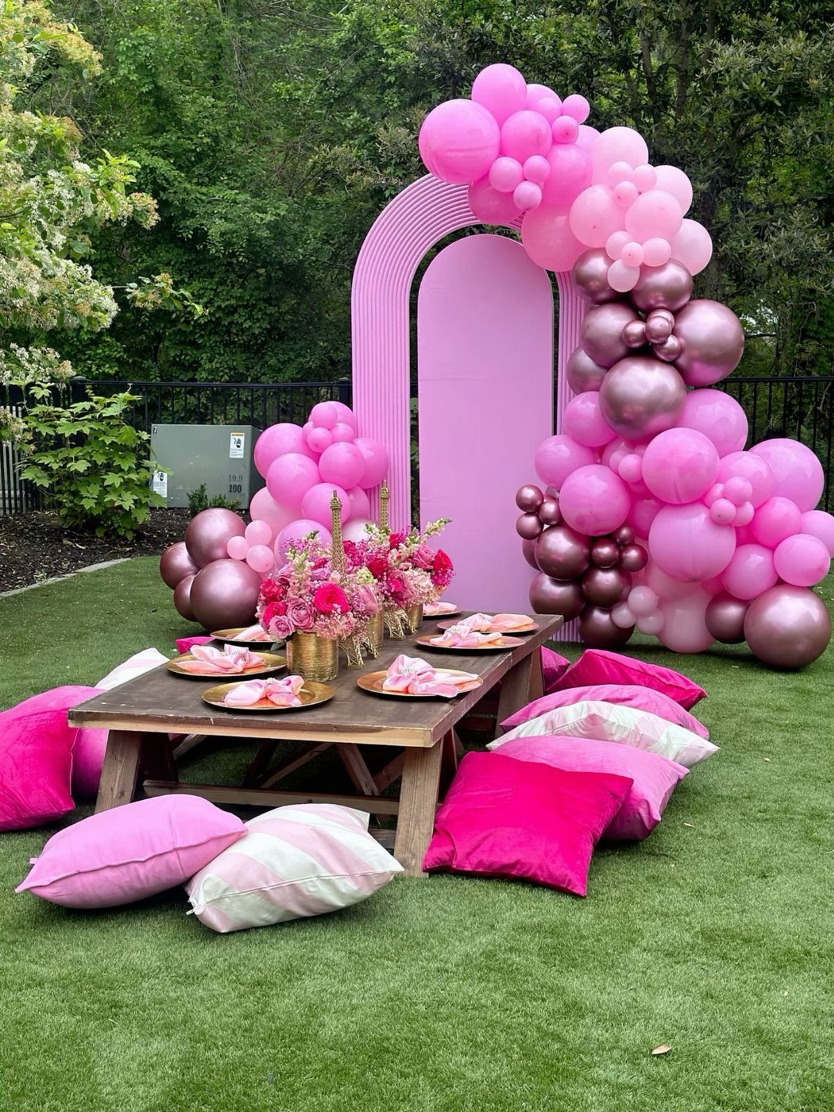 Pink party setup with a balloon arch and low table on green grass.