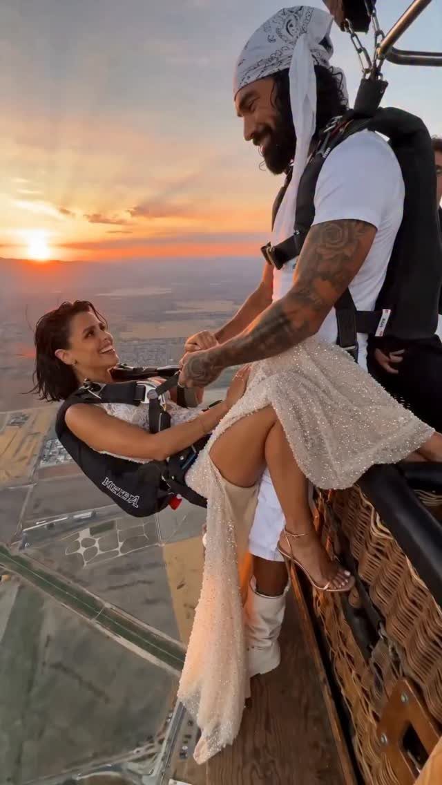 Couple in white clothing posing in a hot air balloon basket at sunset.