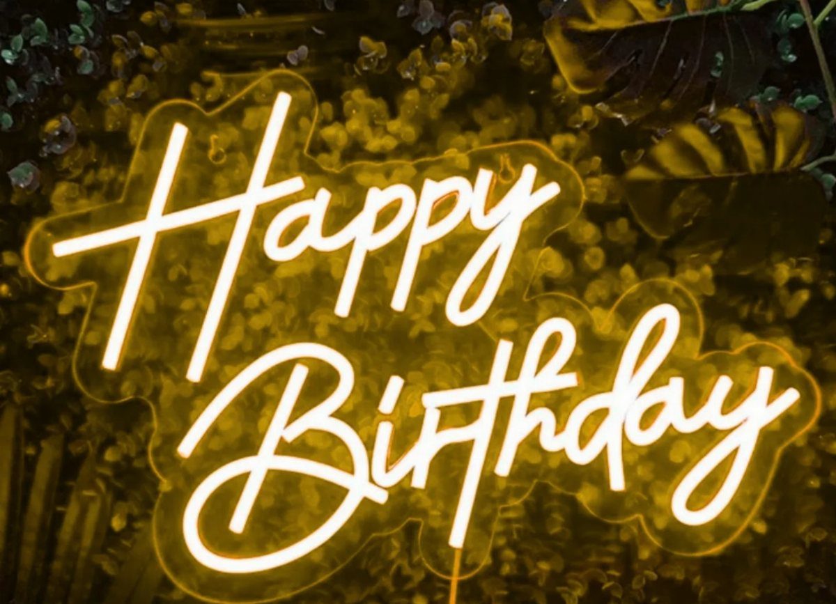 Yellow neon Happy Birthday sign glowing against a dark green leafy background