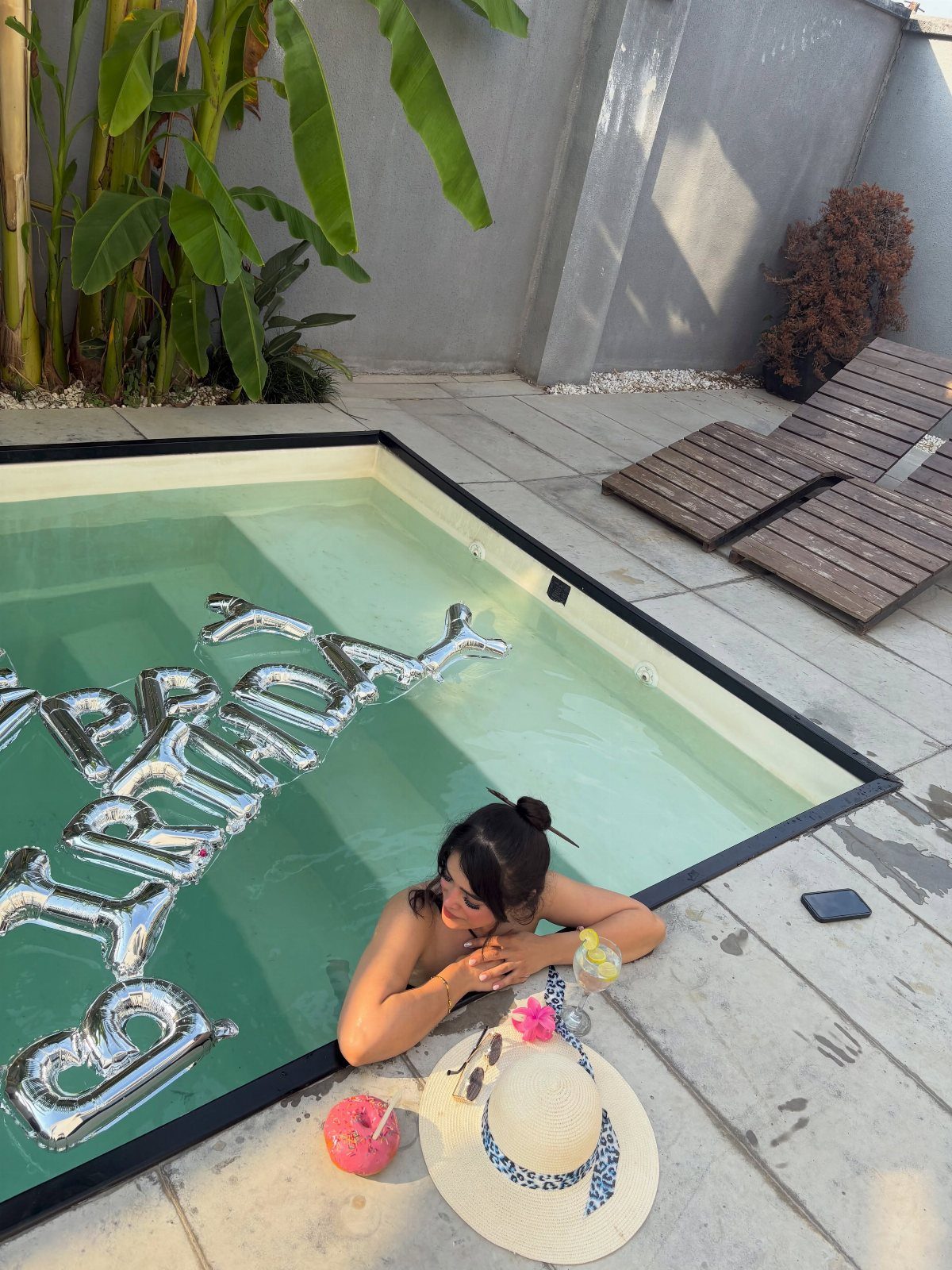 Woman posing in a pool with silver birthday balloons and tropical drinks nearby.