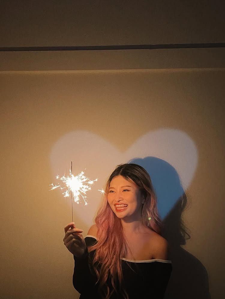 Woman with pink hair holding a lit sparkler against a heart-shaped light projection.