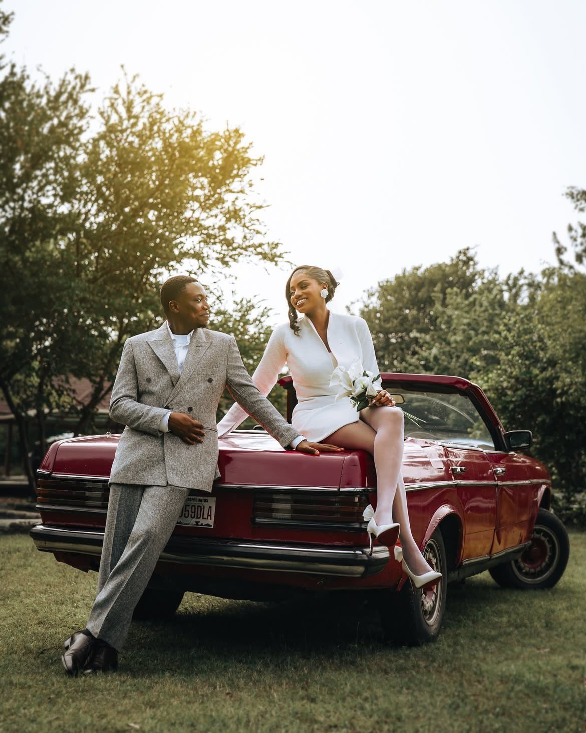Couple posing with a red vintage convertible car in a grassy outdoor setting.