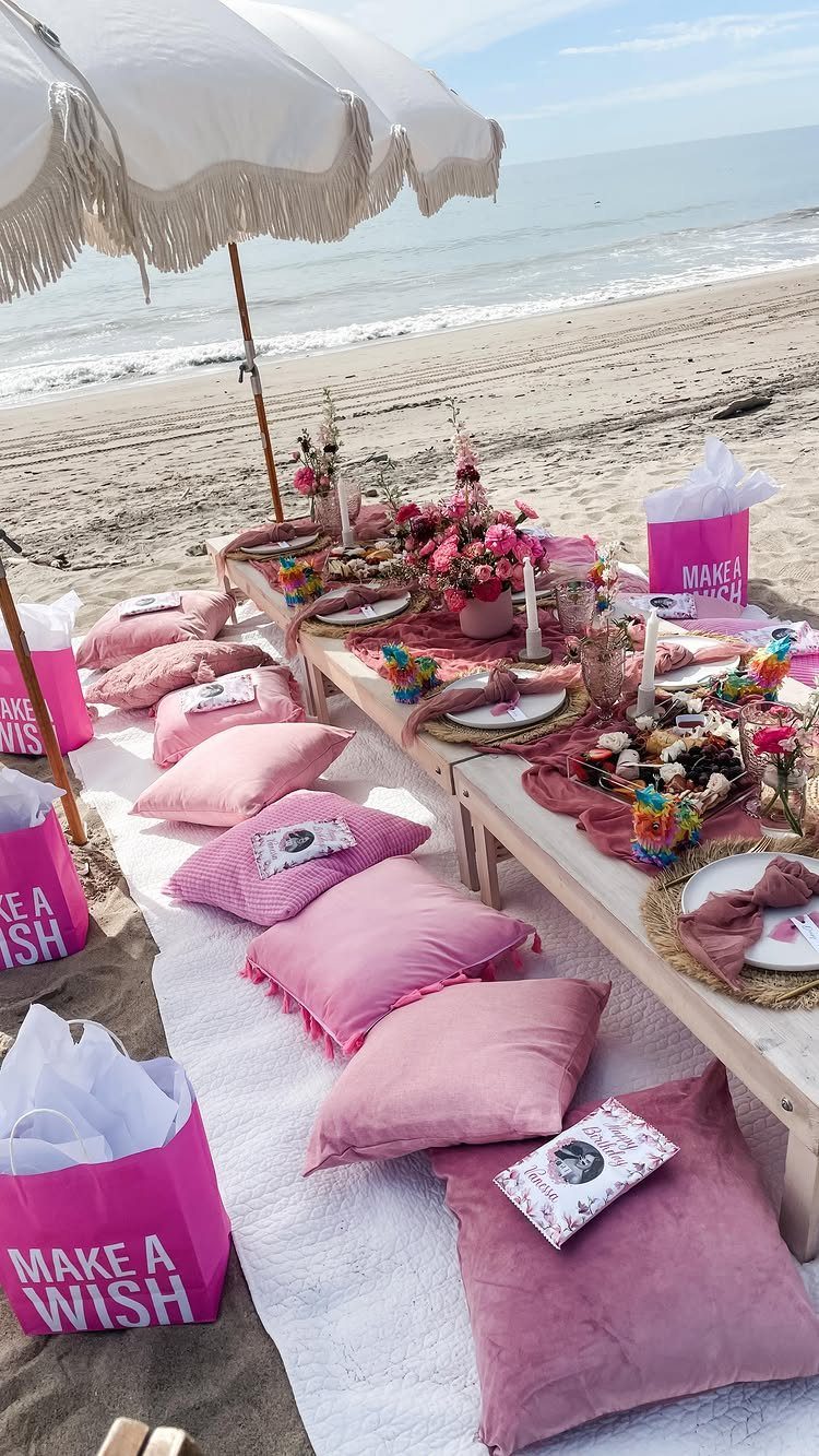 Beach picnic setup with pink pillows, gift bags, and food under a white umbrella.