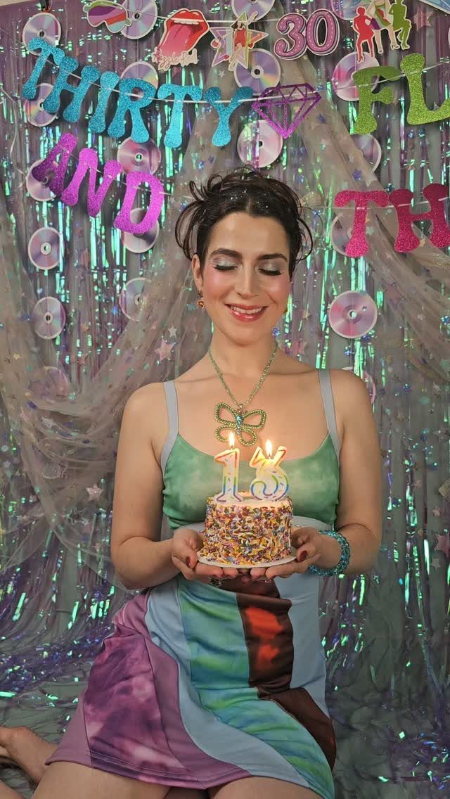 Woman in a colorful dress holding a 30th birthday cake against tinsel backdrop