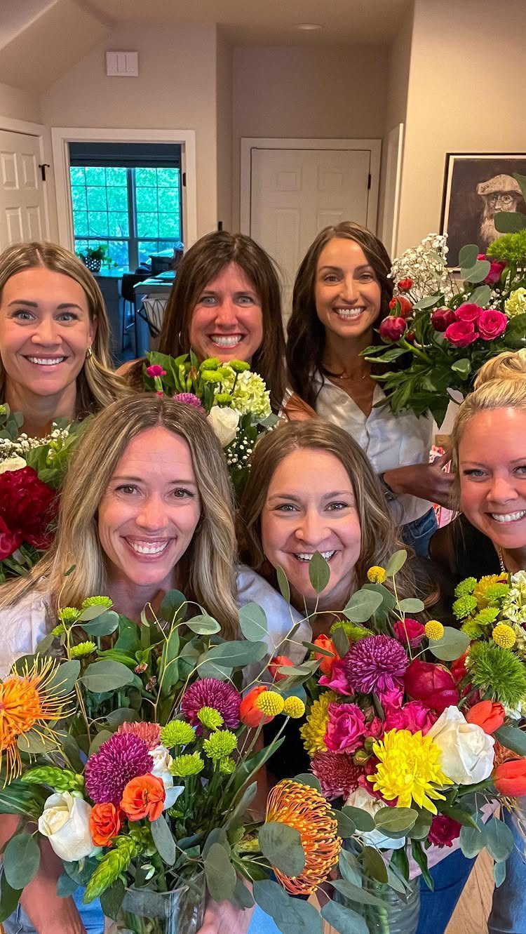 Six smiling women holding colorful floral bouquets in a brightly lit indoor setting.