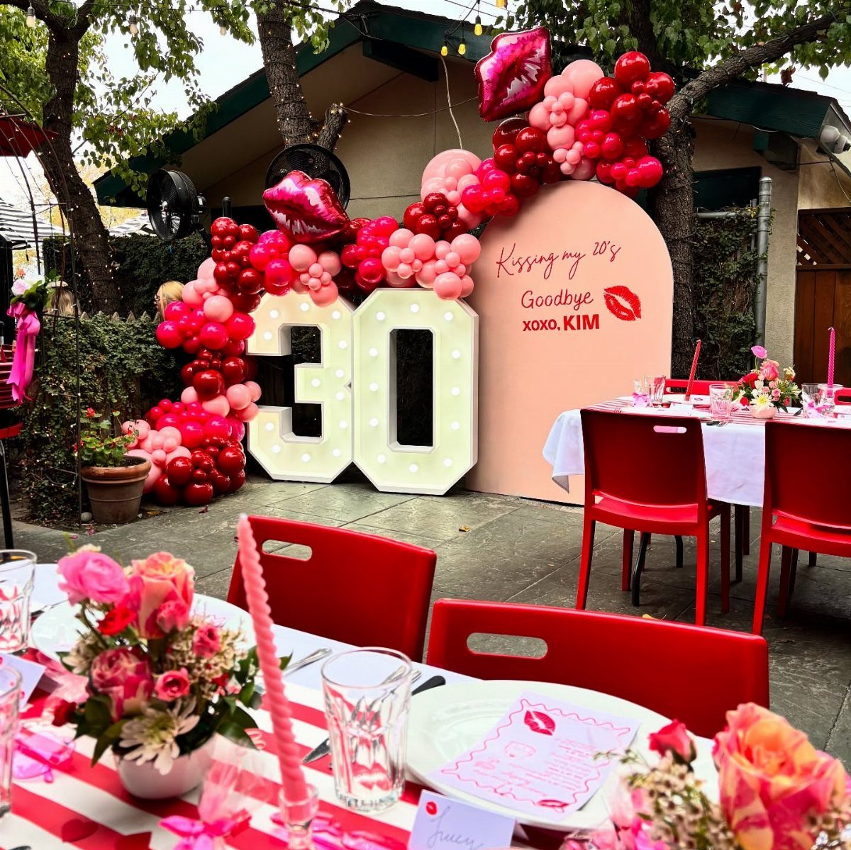 Outdoor 30th birthday party setup with pink balloons, red chairs, and illuminated numbers.