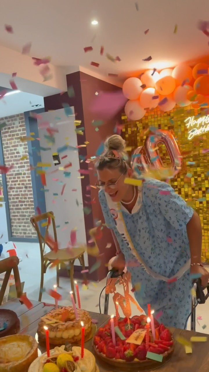 Woman in hospital gown celebrating 40th birthday with cakes, balloons, and confetti.