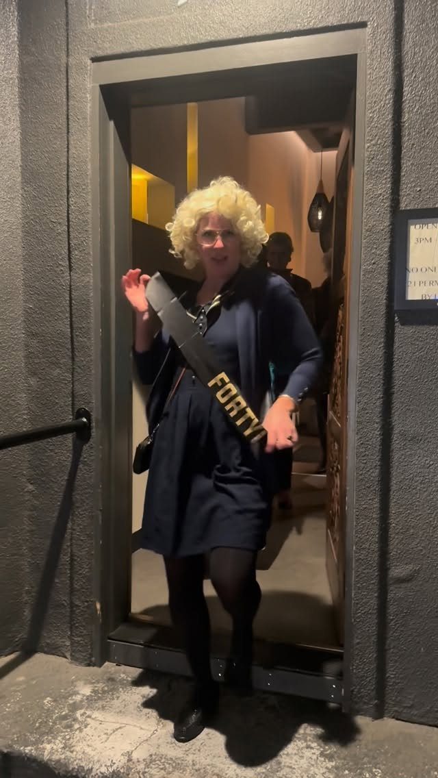 Woman in a blonde wig and 'FORTY' sash posing in a doorway