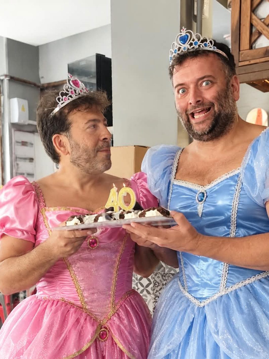 Two bearded men in princess dresses and tiaras holding a 40th birthday cake.